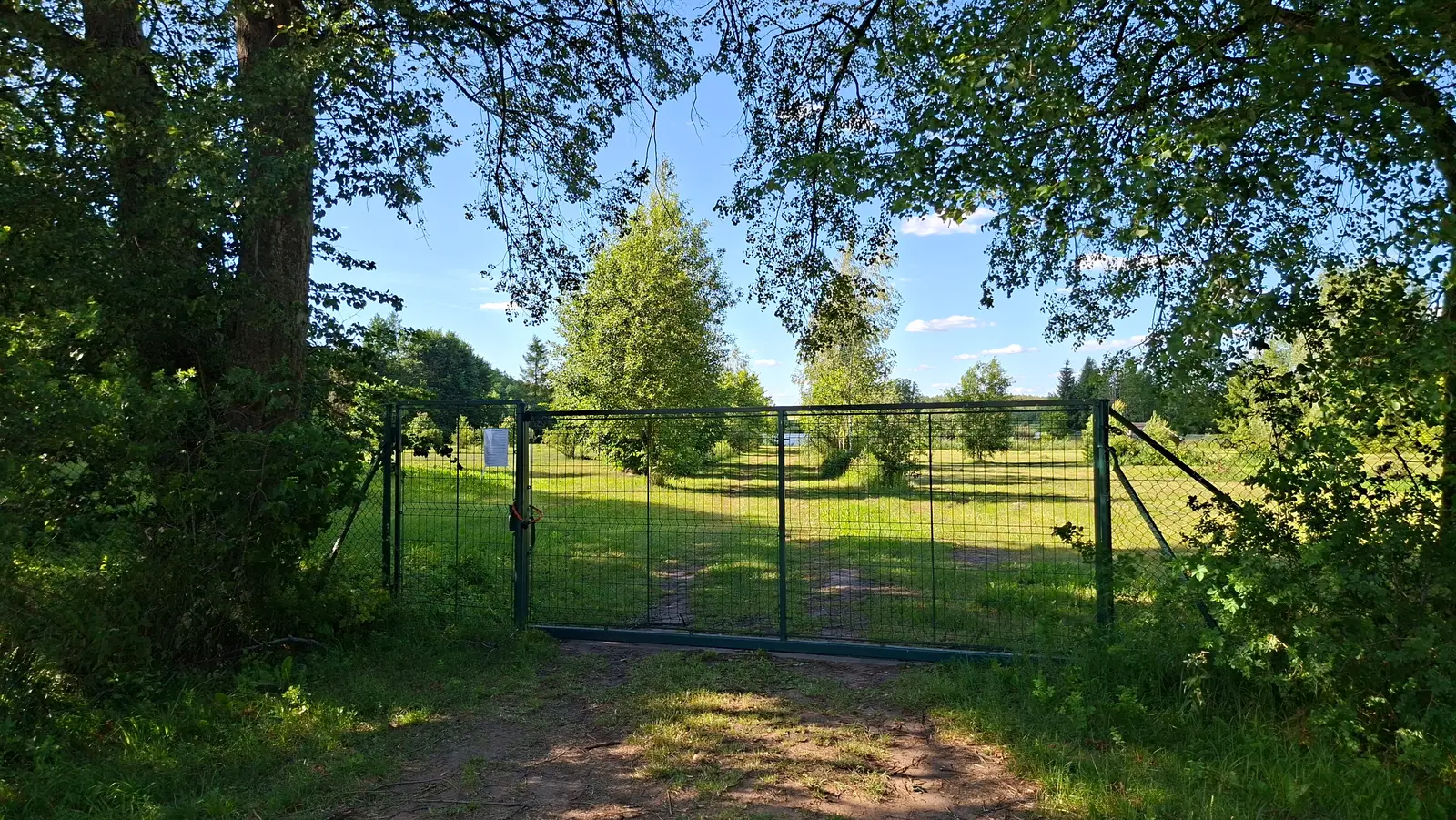 Zdjęcie w formacie poziomym przedstawia metalową, zieloną bramę wjazdową umieszczoną na tle słonecznego, wiejskiego krajobrazu. Brama wykonana jest z siatki drucianej i stanowi główny element kompozycyjny na środku kadru, dzieląc przestrzeń na część zacienioną na pierwszym planie i rozległą, zieloną łąkę w tle. Po lewej stronie bramy widać przytwierdzoną białą tabliczkę z tekstem, natomiast gęste korony drzew po obu stronach kadru tworzą naturalną ramę dla otwartej przestrzeni. Na tabliczce widoczny jest następujący tekst: „UWAGA! TEREN PRYWATNY. WSTĘP WZBRONIONY. OSOBY NIEUPOWAŻNIONE BĘDĄ ŚCIGANE. WŁAŚCICIEL TERENU”. Całość utrzymana jest w jasnej, letniej kolorystyce z wyraźnym zróżnicowaniem między światłem słonecznym a cieniem rzucanym przez bujną roślinność.